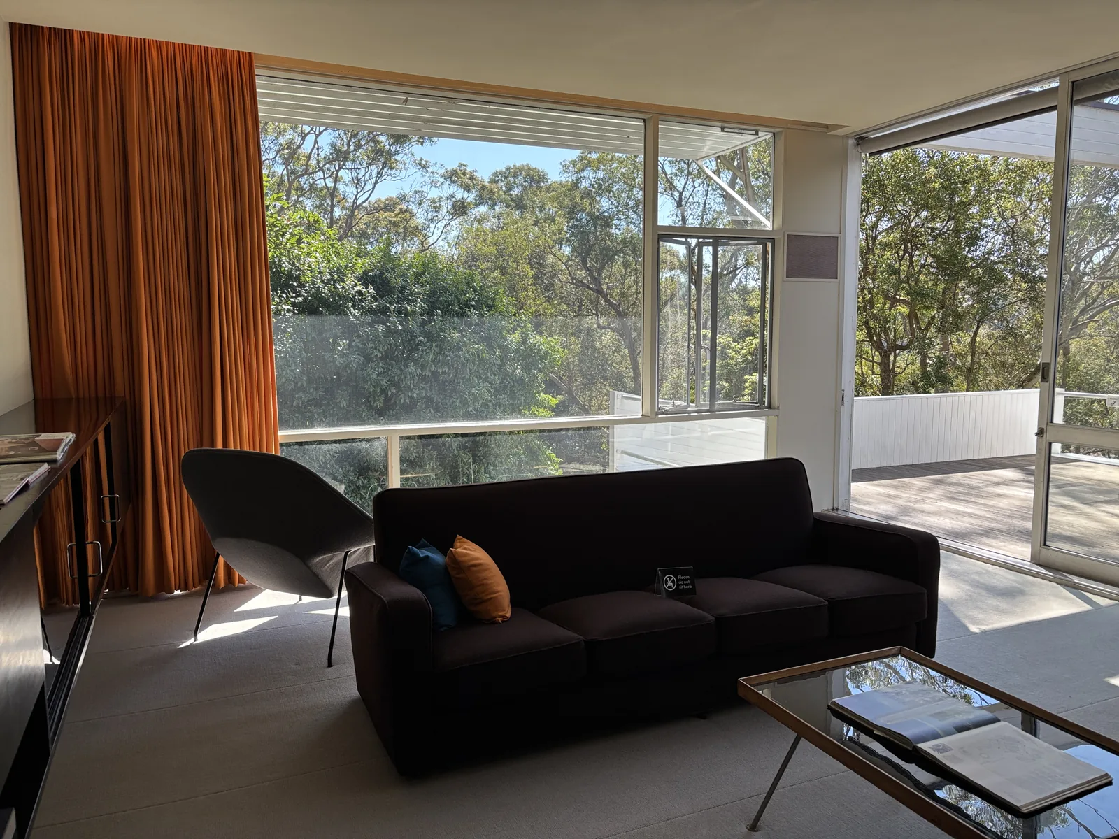 Living room with a sofa and chair beside broad windows overlooking trees, and a glazed opening towards a terrace.