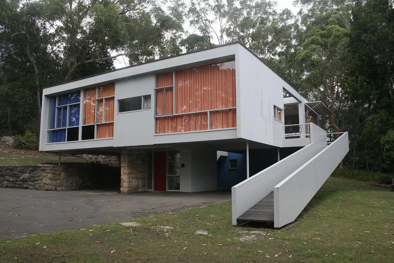 Rose Seidler House among trees, with broad upper-level windows, a stone-faced lower wall and an external ramp rising to a landing.